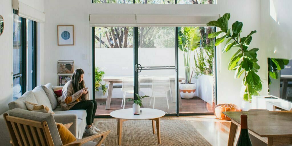 A wide image of a loungeroom and a garden outside. A woman is seated on a couch typing on a mobile phone.